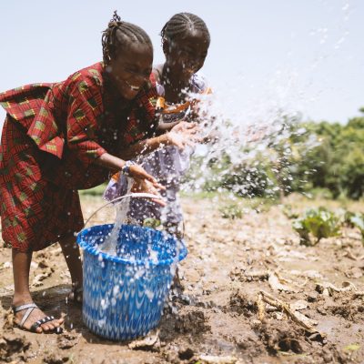 Two playful little African sisters irrigating their father's cabbage field, with eyes closed and a wide smile on their faces